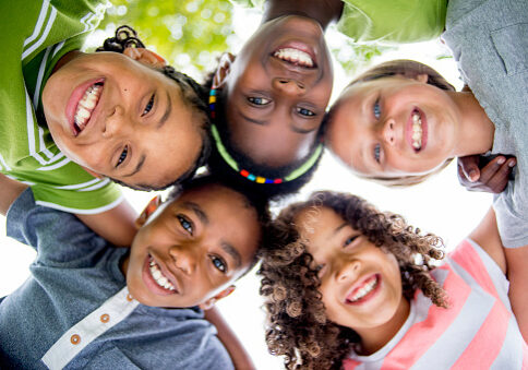 A multi-ethnic group of elementary age children are huddled together, standing in a circle with their heads in the center looking down at the camera. They are smiling and looking at the camera.